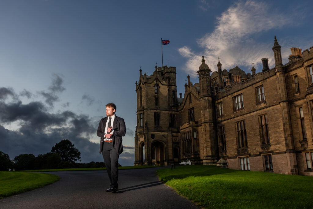 A man in a suit walks on a path near a large stone building with a flag on top, under a partly cloudy sky at sundown.