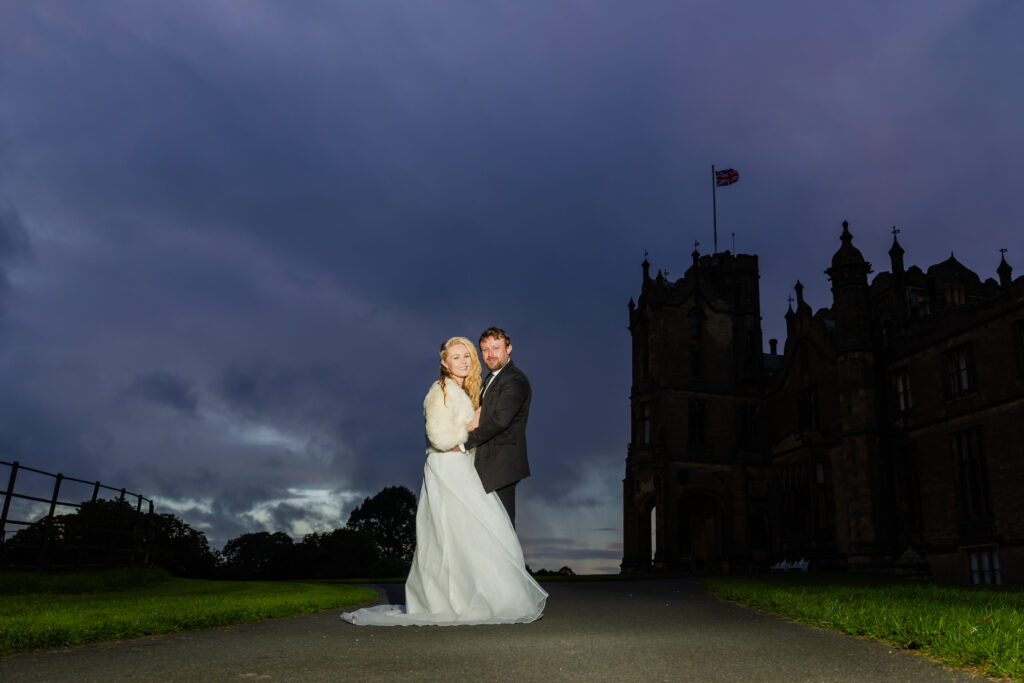 A bride and groom stand together on a path outside a large historic building at dusk, with a dramatic cloudy sky in the background.