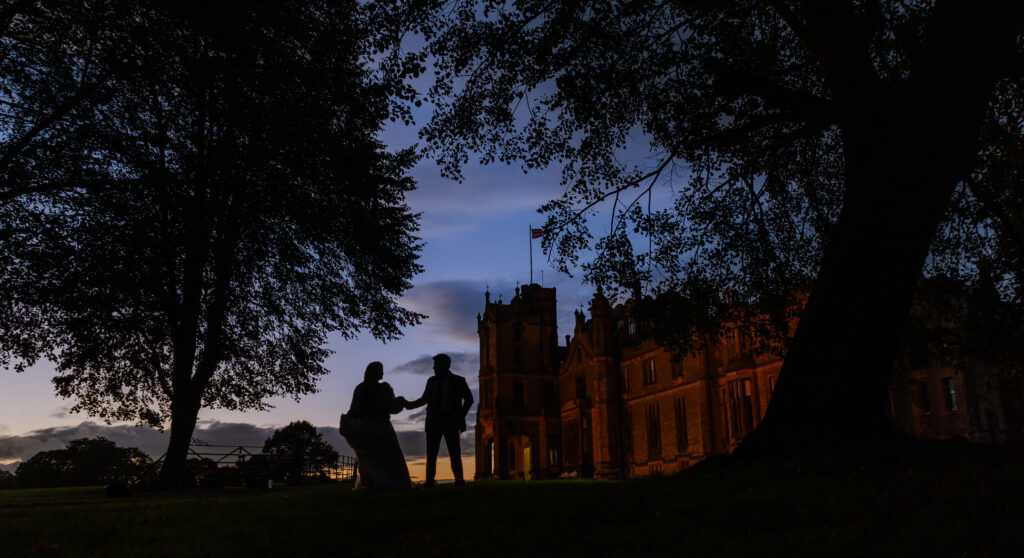 Silhouette of a couple holding hands near a large historic building at dusk, with trees framing the scene and a blue and orange sky in the background.