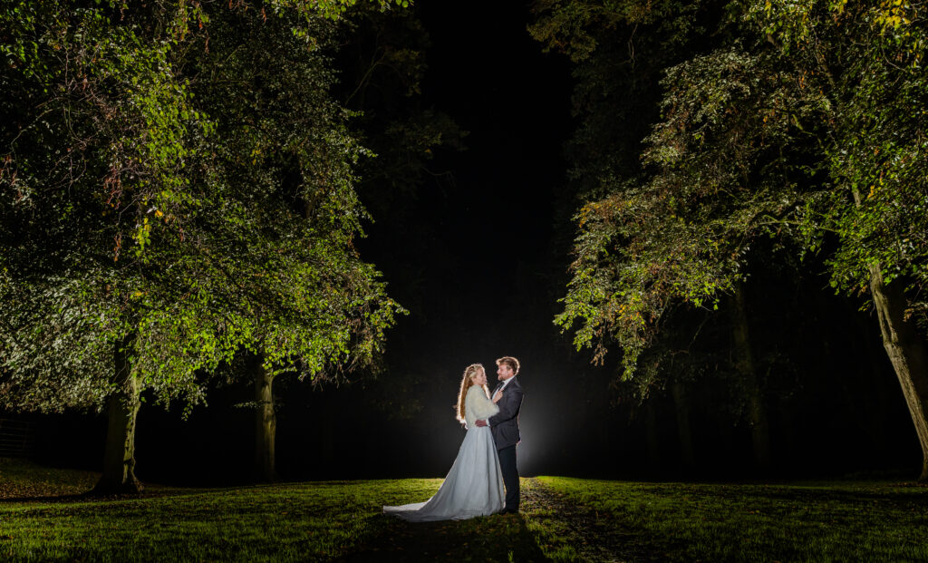 A bride and groom stand facing each other on grass at night, surrounded by tall trees with bright light illuminating them from behind.