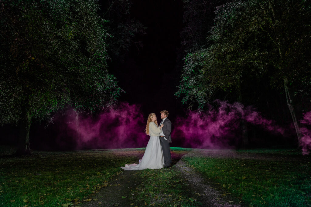A bride and groom stand together on a path at night, surrounded by trees and illuminated by purple-pink coloured smoke in the background.