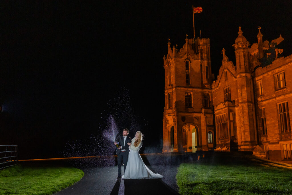 A bride and groom stand on a path at night outside a lit castle, with the groom spraying a bottle, creating a burst of mist in the air.