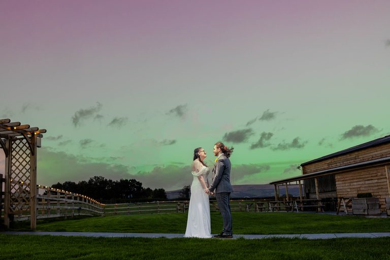 A bride in a white wedding dress and a groom in a grey suit holding hands and looking at each other outdoors at dusk.